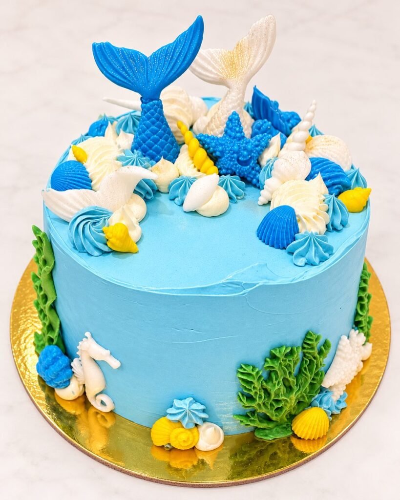 Sky-blue buttercream cake topped with blue and white mermaid tails, a starfish and seashells, with seaweed and a seahorse around the base