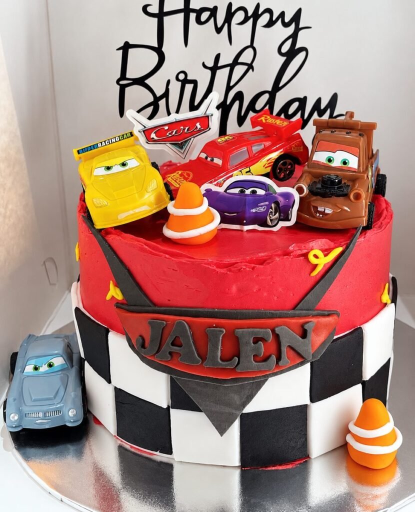 Red and chequered-flag birthday cake topped with Cars toy racing cars and traffic cones