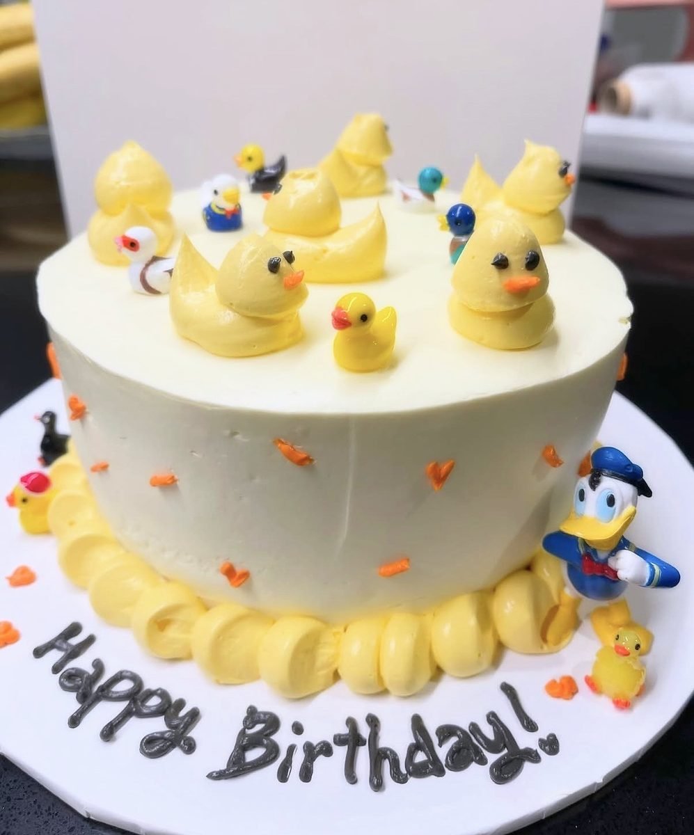 White buttercream cake topped with piped yellow ducklings and small rubber duck figurines, with Happy Birthday lettering