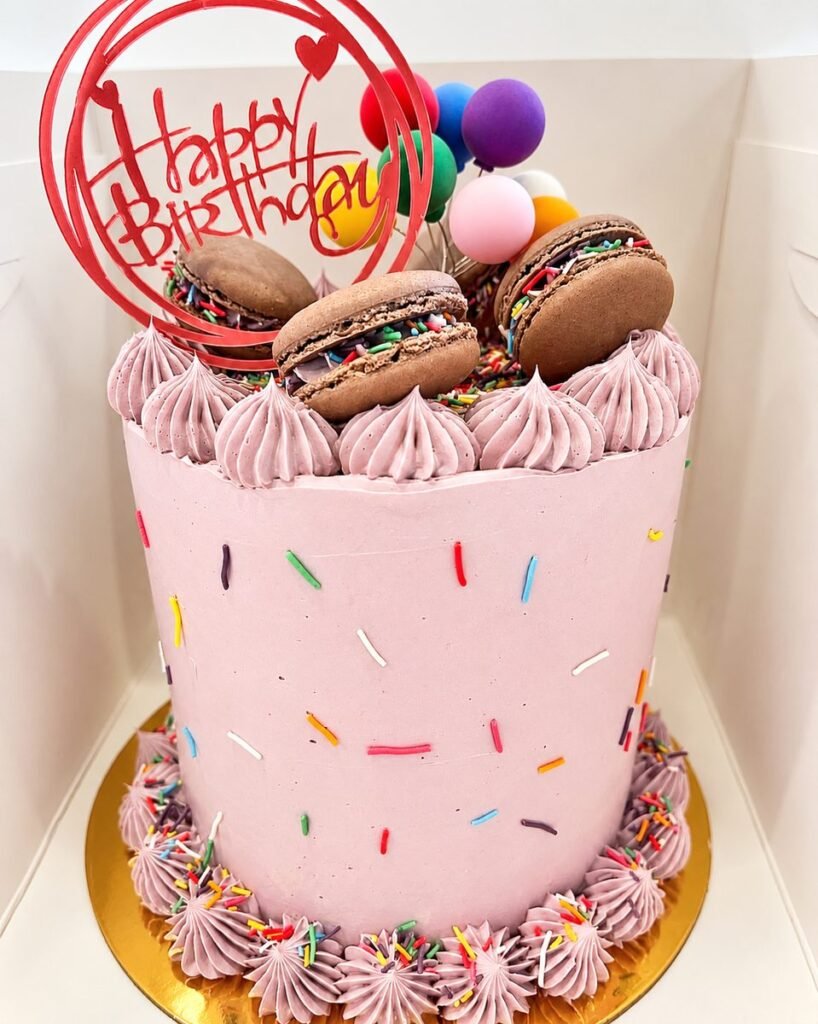 Pink buttercream birthday cake covered in sprinkles and topped with chocolate macarons
