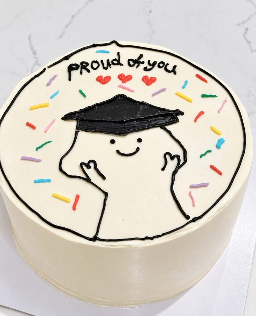 White lunchbox-style cake piped with a smiling figure in a graduation cap, hearts, sprinkles and the words proud of you